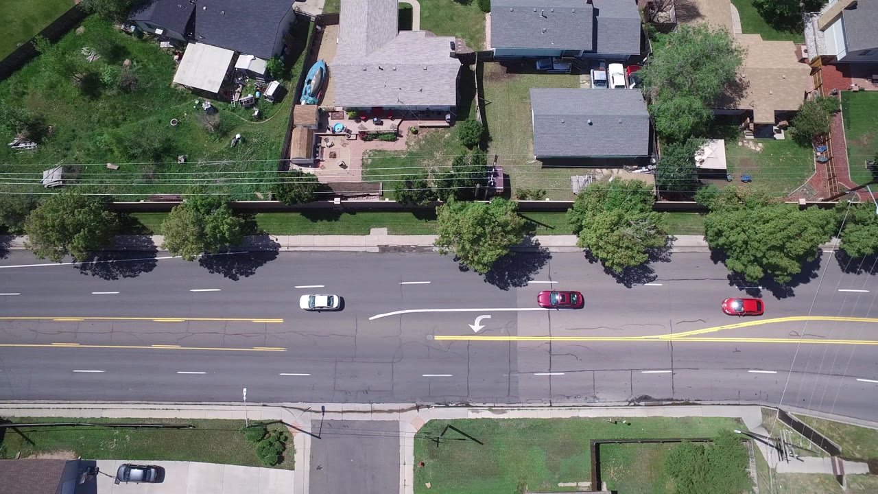 Overhead Westminster, Colorado frame with white, red, and dark cars on a broad residential road bordered by yards, trees, sidewalks, and house roofs, with no visible people.