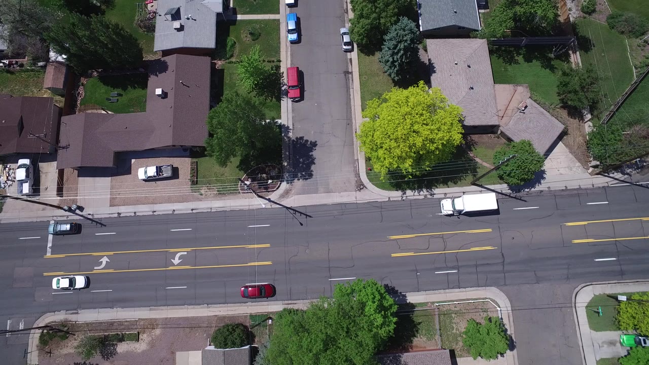 Overhead frame from Westminster, Colorado showing houses, lawns, mature trees, parked cars, a multi-lane road with yellow lane markings, and no visible people.