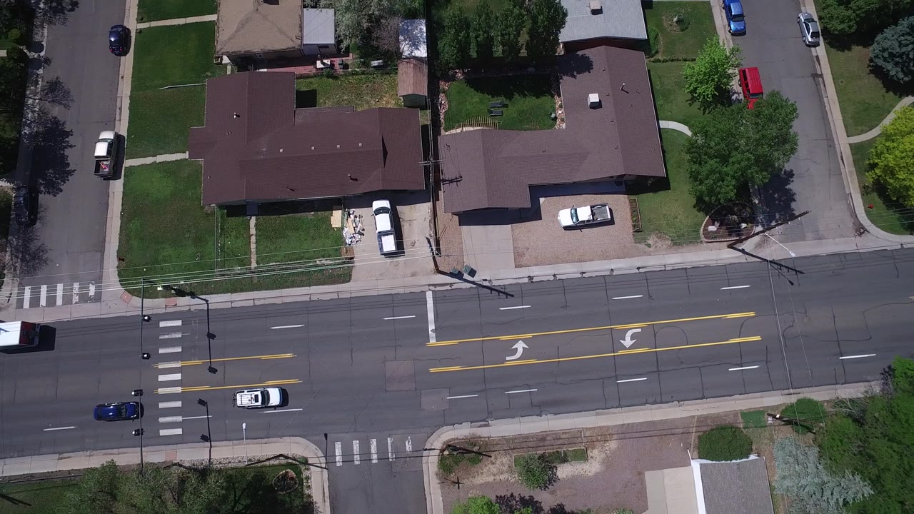 Top-down Westminster, Colorado view with two homes near the center, a broad road across the lower frame, vehicles on the lanes, and no visible people.