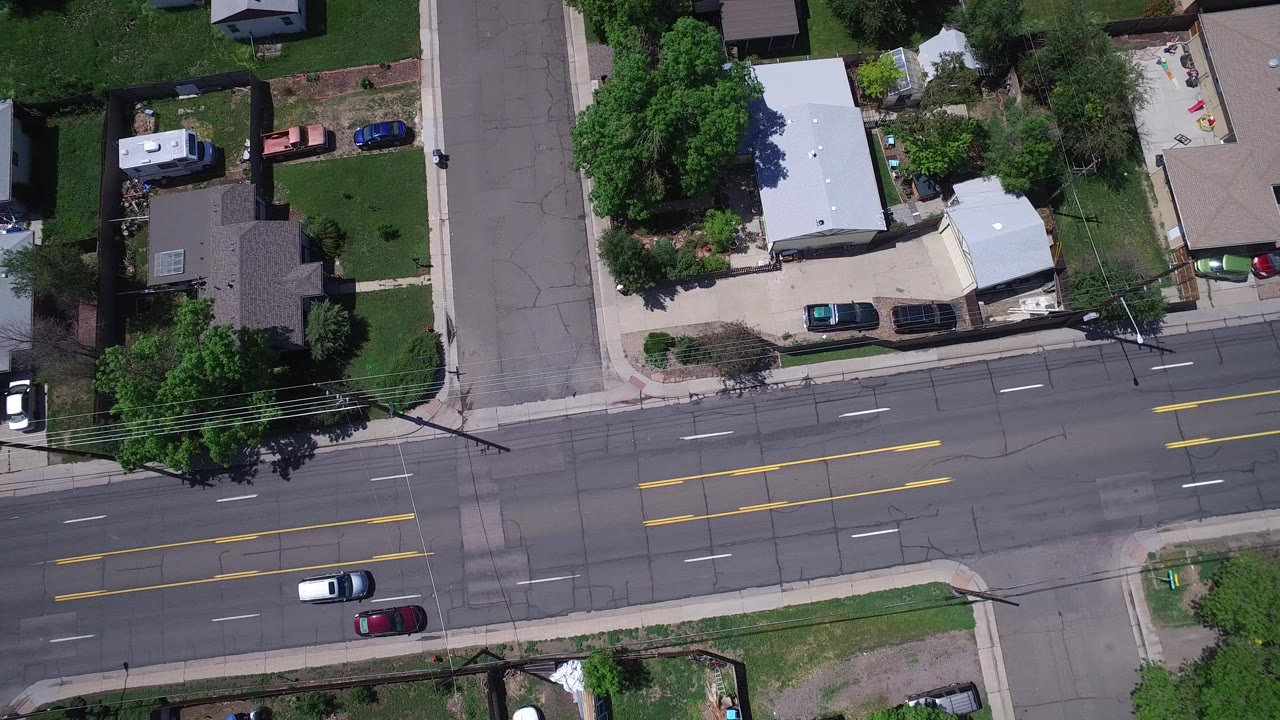 Aerial view in Westminster, Colorado with the main road running across frame, a side street extending upward, rooftops, yards, trees, sidewalks, and no clearly visible people.