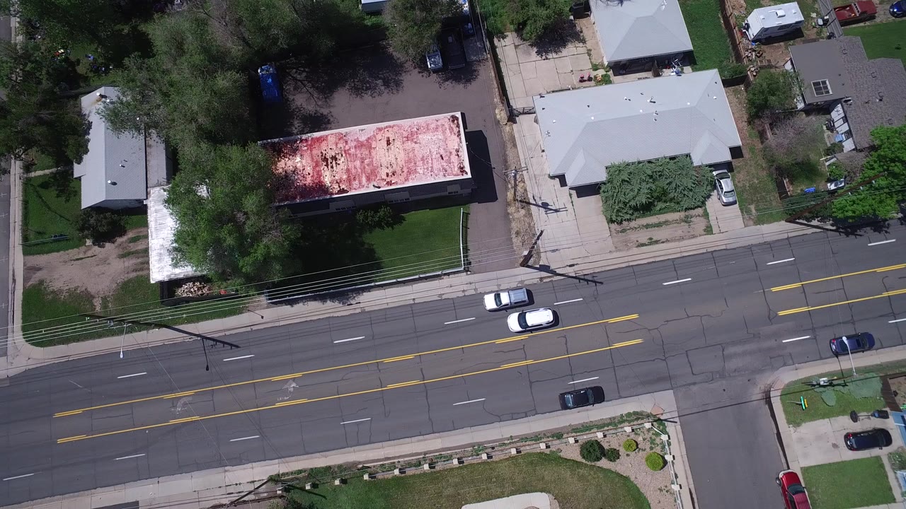 Overhead view of a suburban road with cars, trees, lawns, and low buildings.