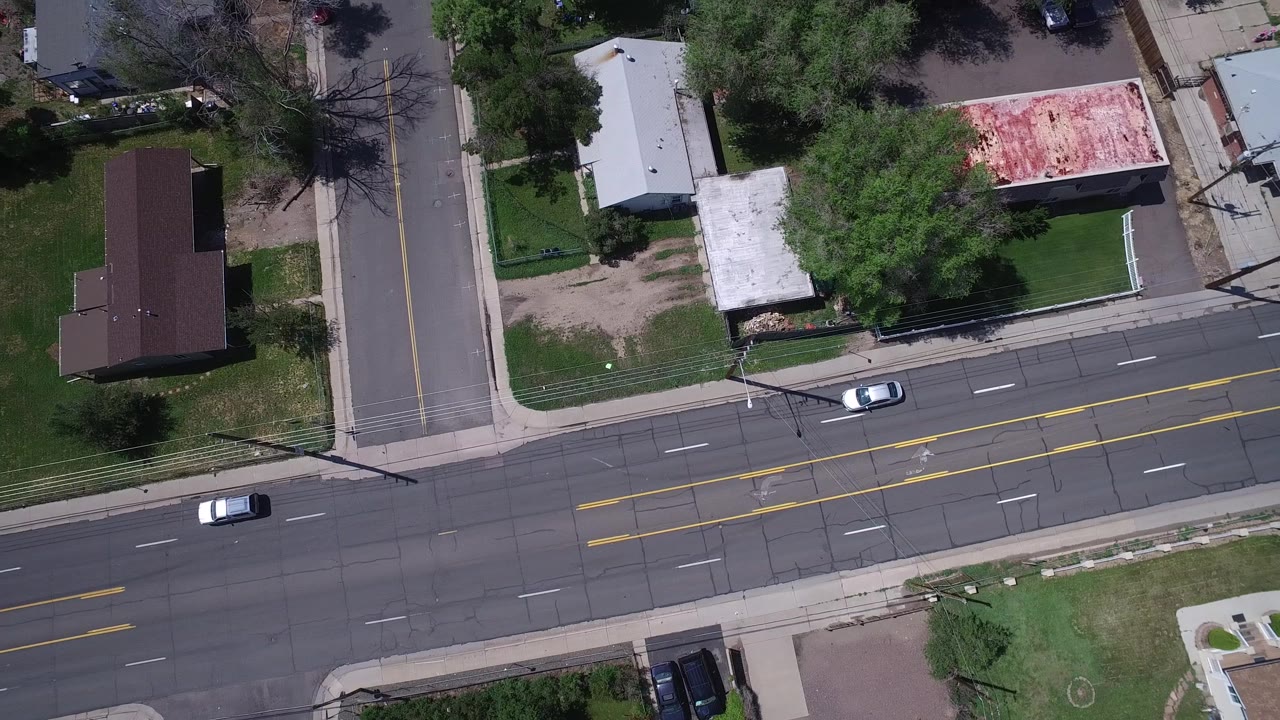 Aerial frame over Westminster, Colorado showing sparse cars on a wide road, a side street, green lawns, rooftops, trees, and no visible people.