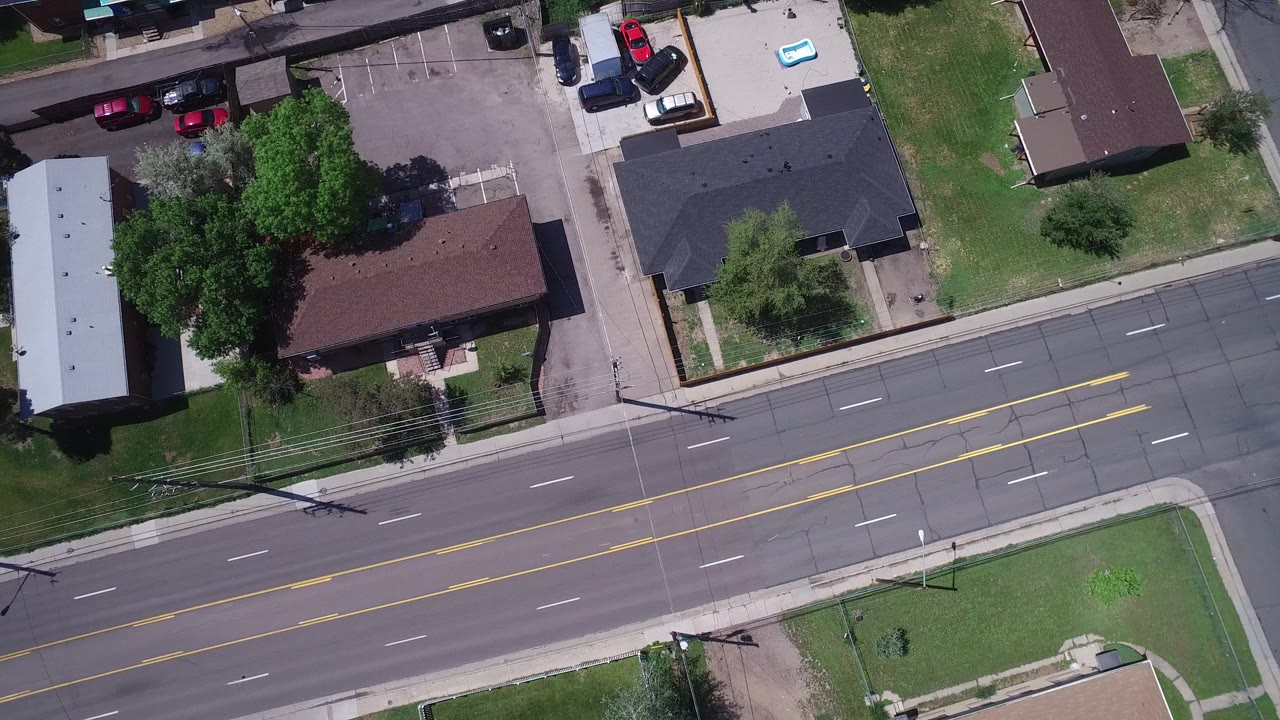 Aerial frame from Westminster, Colorado with the main road centered lower in view, a side street at right, low buildings, driveways, trees, parked vehicles, and no visible people.