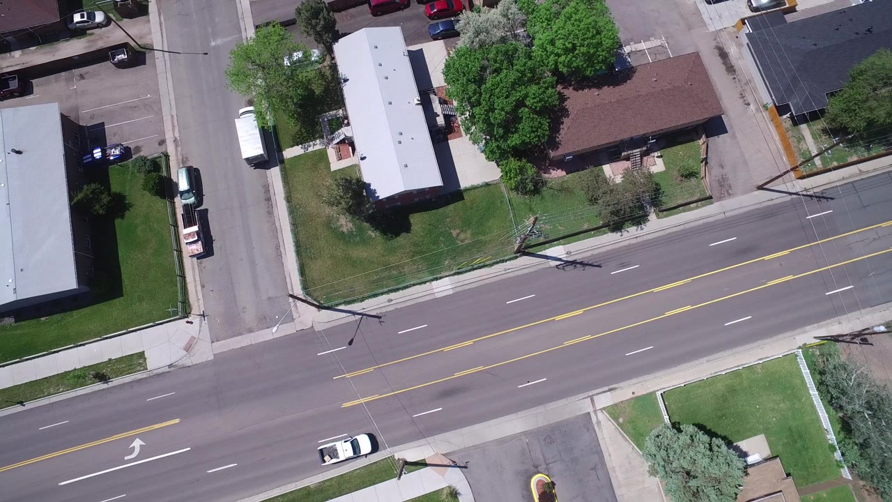 Overhead view of a wide street, side road, buildings, lawns, trees, and a white car.