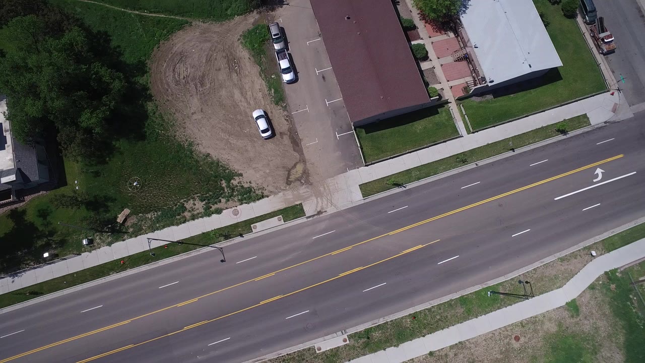 Overhead view of a multi-lane road beside trees, sidewalks, parked cars, and low buildings.