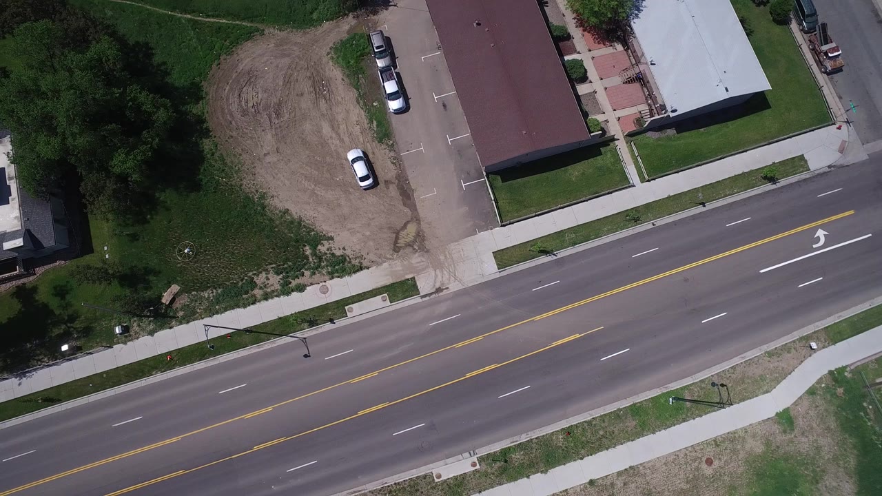 Aerial view in Westminster, Colorado with the roadway shifted across frame, yellow markings centered, nearby sidewalks, parked cars, trees, and no visible people.
