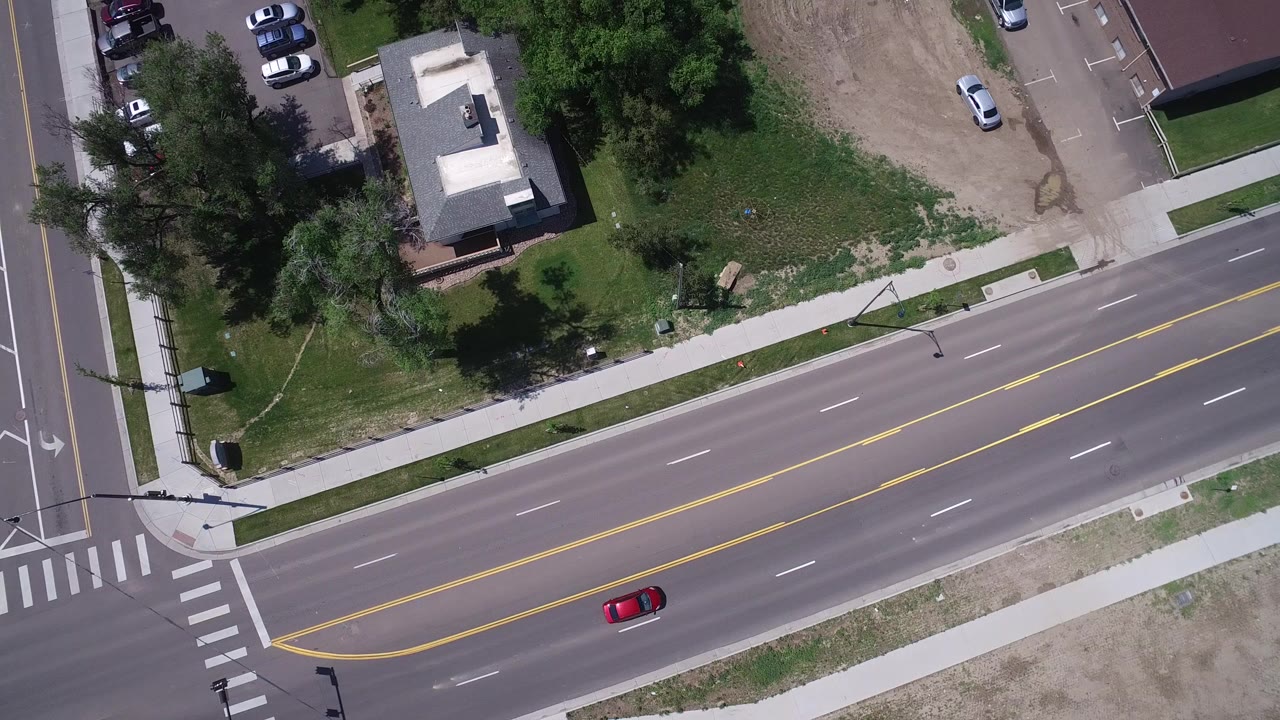 High aerial view in Westminster, Colorado showing a red car near a multi-lane intersection, crosswalks, sidewalks, lawns, trees, and small buildings with no visible people.