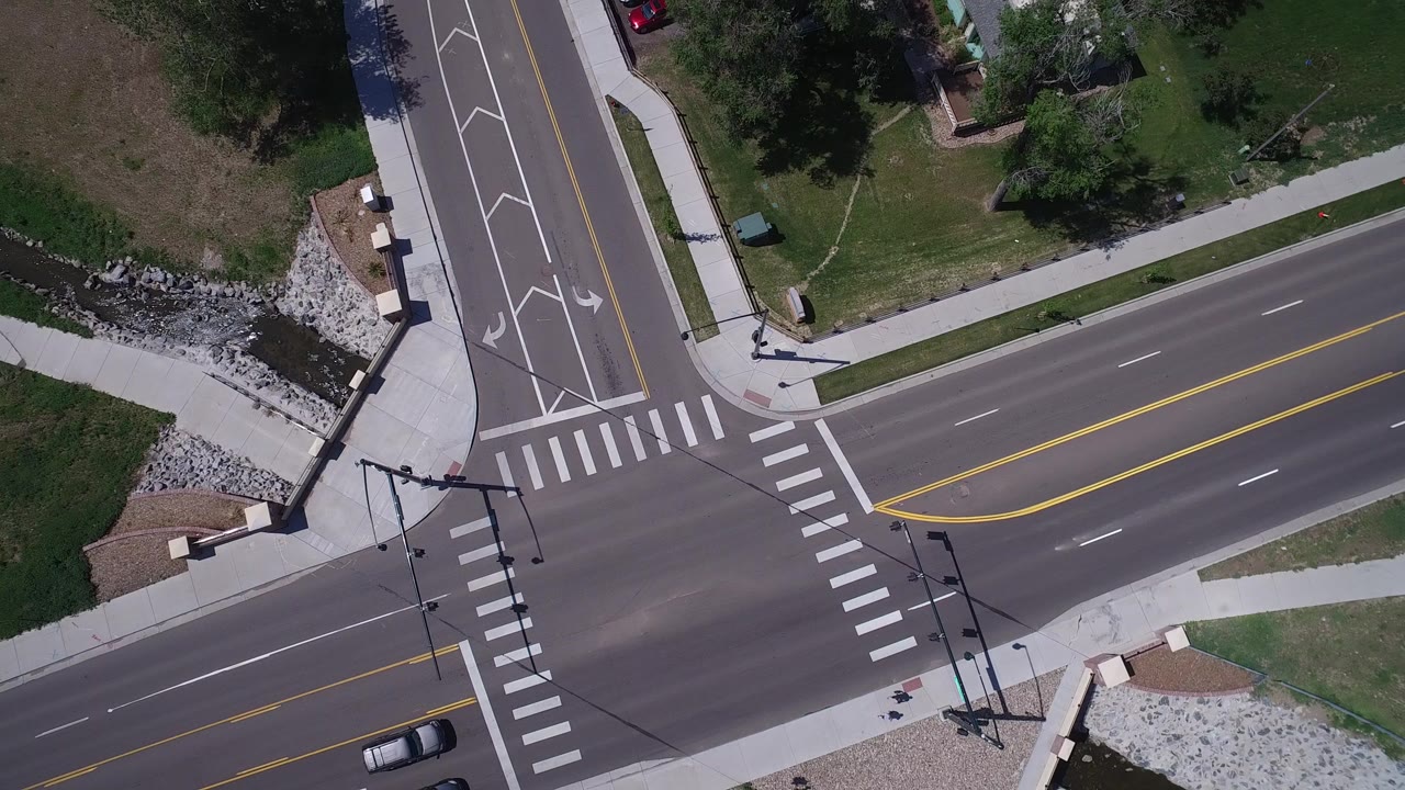 Overhead frame of a Westminster, Colorado T-shaped junction with striped crosswalks, turn arrows, sidewalks, grass, a canal edge, and no visible people.
