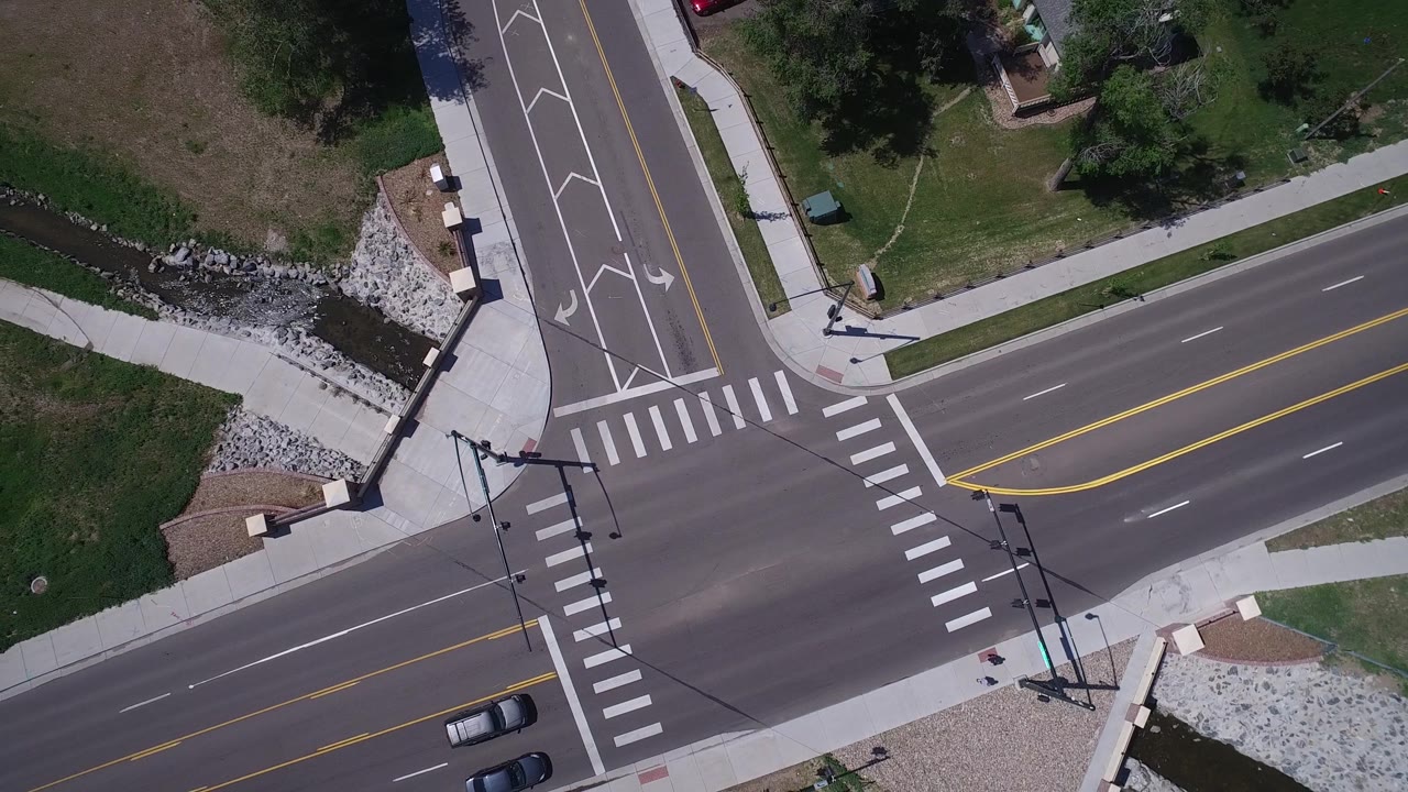 Aerial view of a paved T-junction with lane arrows, crosswalks, grass, and drainage channel.