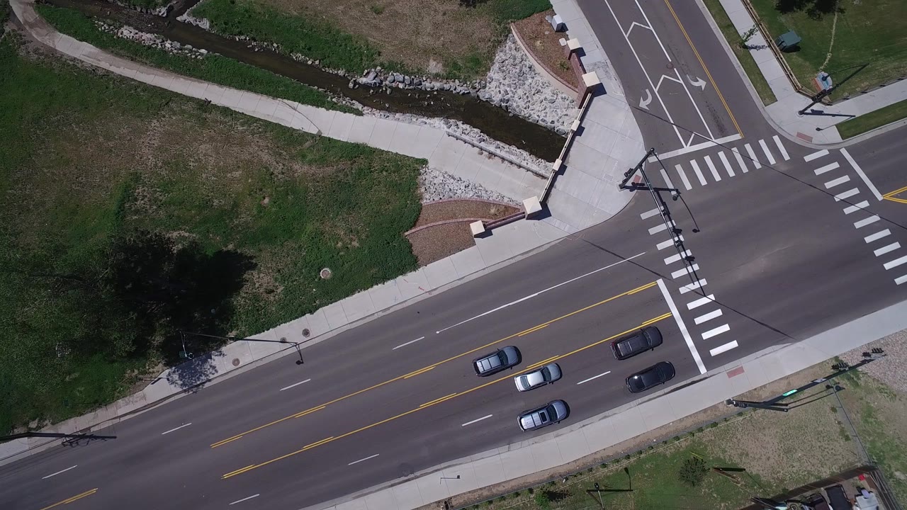 Aerial Westminster, Colorado frame with several cars shifted along the road lanes near the intersection crosswalks, bordered by grass, sidewalks, and a narrow channel, with no visible people.