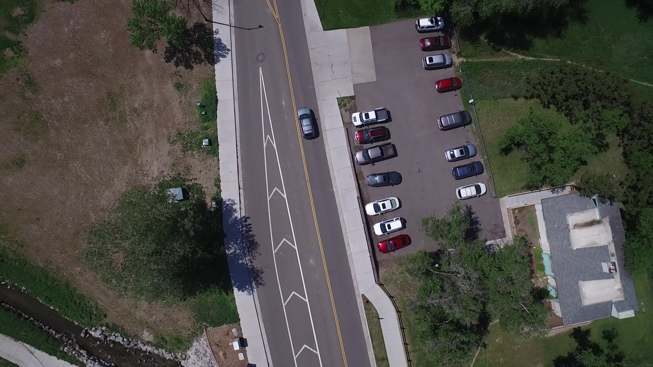 Overhead view of road, parking lot, cars, grass, trees, and a small building.
