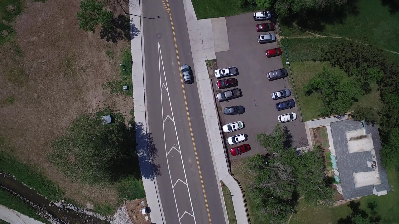 A second overhead frame in Westminster, Colorado keeps the road on the left and shows more of the parking rows, brown and green grass, trees, and the small building, with no visible people.
