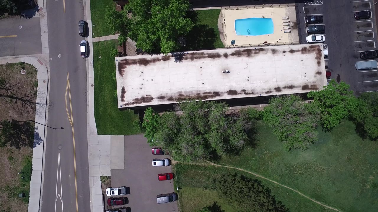 Aerial frame over Westminster, Colorado with the blue pool near the upper center, a long pale roof below it, tree shade across the grass, parked cars, and a road along the left side, with no clearly identifiable people.