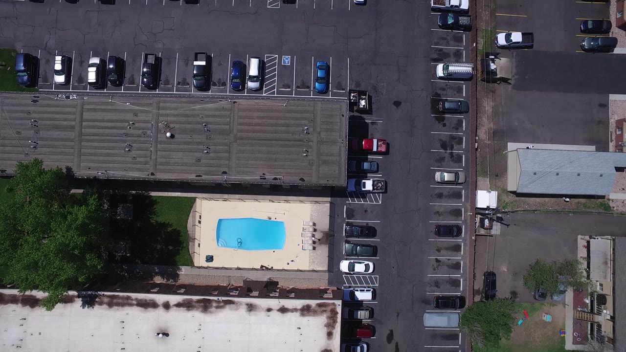 Overhead Westminster, Colorado view with the pool shifted lower in frame, parked vehicles along marked spaces, building roofs, a green tree canopy, and no visible people.