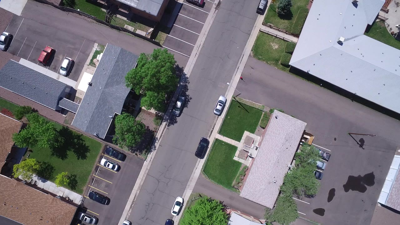 Overhead suburban street with rooftops, trees, lawns, and parked cars.