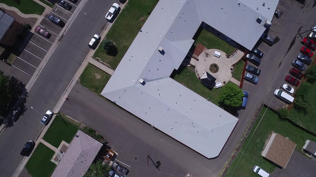 Top-down aerial frame in Westminster, Colorado showing the corner of a gray-roofed building, adjacent streets, sidewalks, lawns, parked cars, and no visible people.