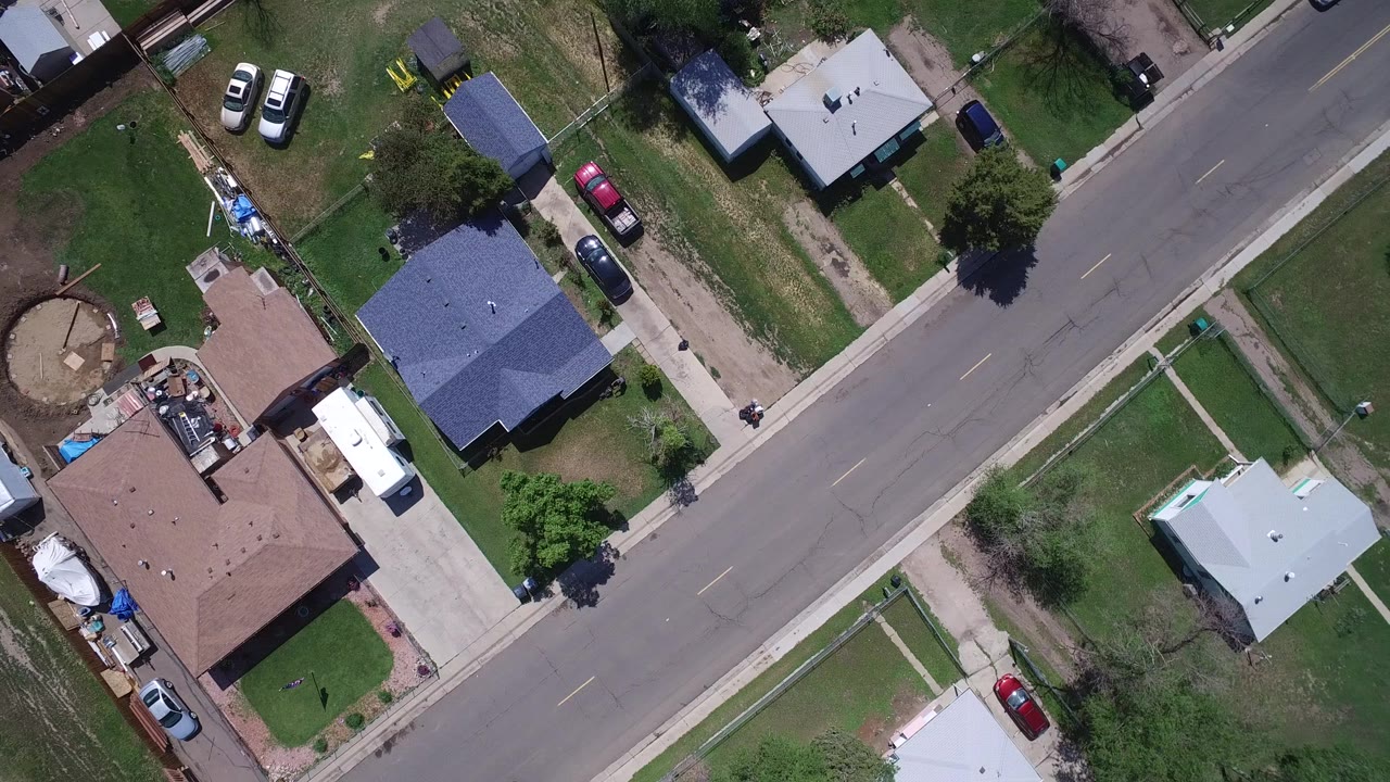 Aerial frame over Westminster, Colorado with the road running diagonally between houses, open lawns, a driveway, parked vehicles, and no visible people.