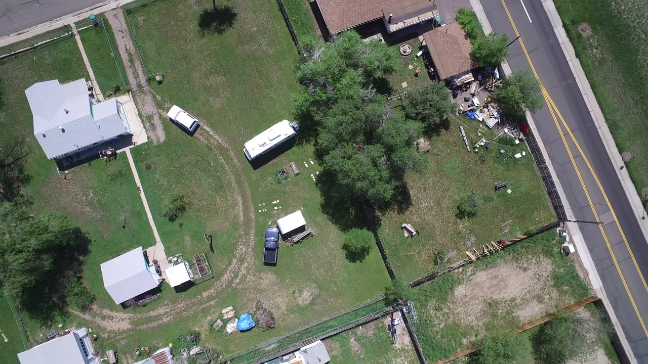Overhead frame of Westminster, Colorado showing residential lawns, small houses, trees, a white trailer or RV, parked vehicles, and a two-lane road at the right with no visible people.