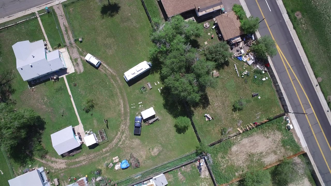 Aerial view over Westminster, Colorado with the road running along the right edge, lawns and trees in the center, houses at the upper edge, parked vehicles, and no visible people.