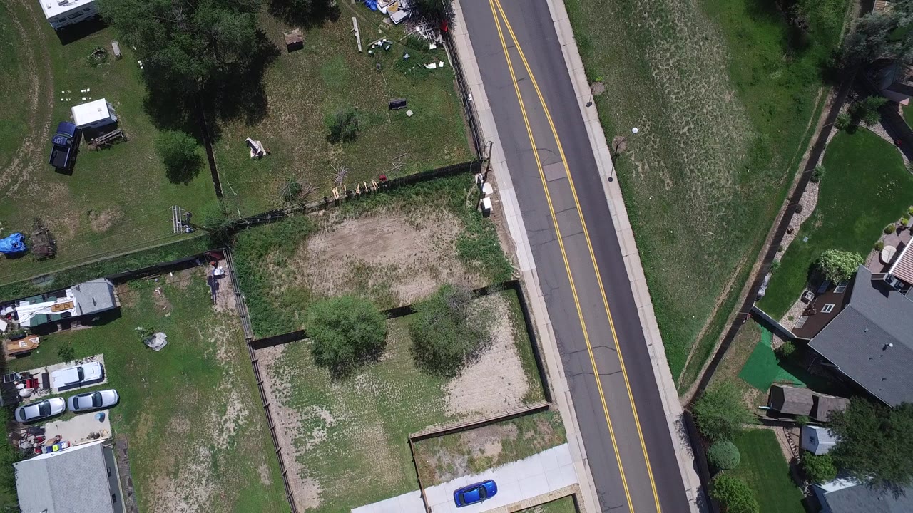 High overhead view in Westminster, Colorado of a two-lane road beside fenced yards, lawns, trees, parked vehicles, and residential roofs, with no visible people.