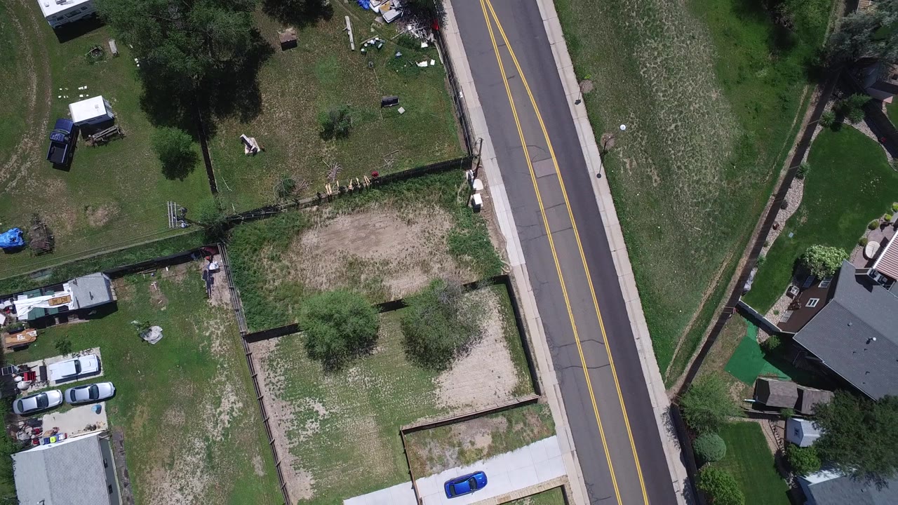 Westminster, Colorado aerial frame showing a vertical neighborhood road, green and patchy lawns, backyard fencing, several rooftops, parked cars, and no visible people.