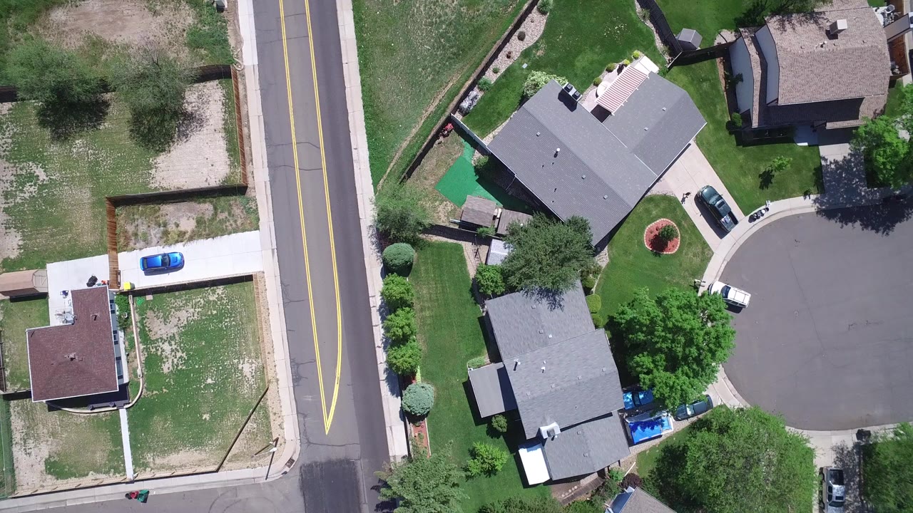Aerial frame over Westminster, Colorado showing a two-lane road beside private homes, driveways, lawns, trees, and parked cars, with no clearly identifiable people visible.