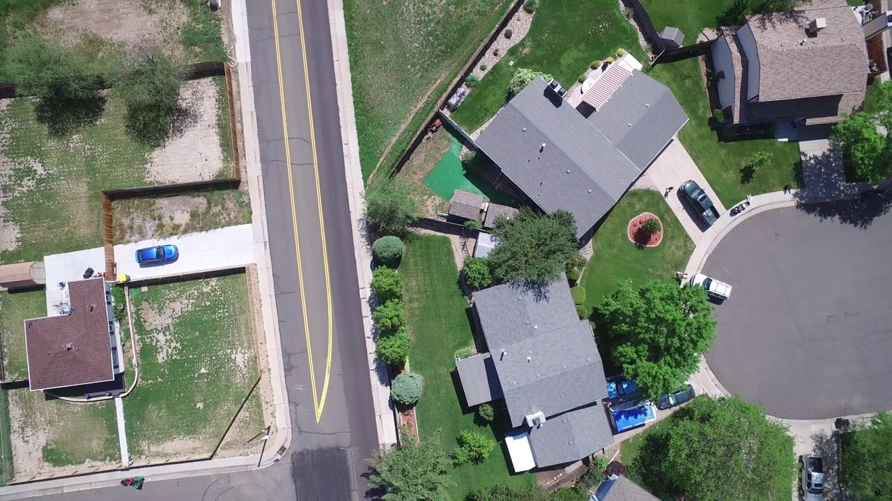 Aerial view in Westminster, Colorado with the main road running vertically beside roofs, yards, a partial cul-de-sac, and several parked vehicles in bright sun.