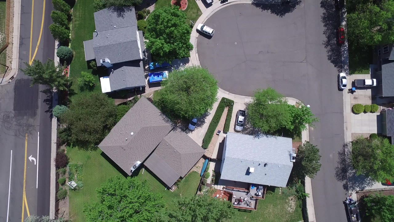 Overhead Westminster, Colorado frame showing two main homes, leafy yards, driveways, parked vehicles, and a residential intersection with no visible people.