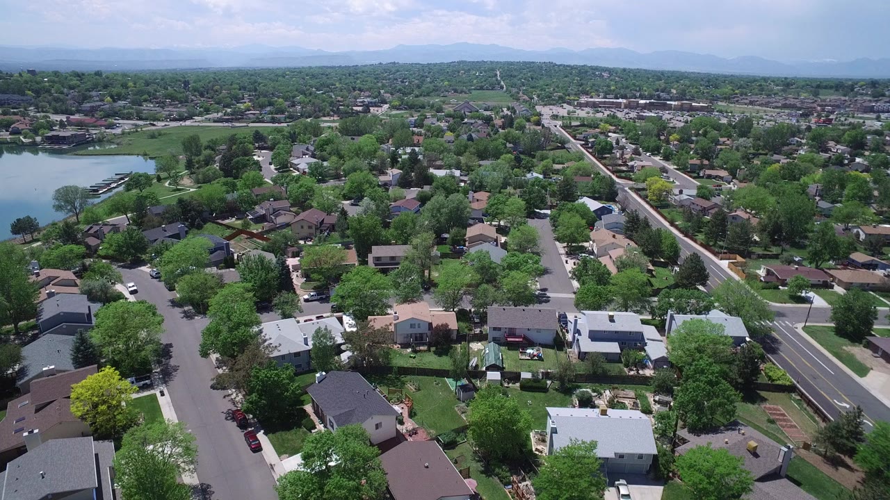 Aerial view over suburban homes, trees, streets, and a lake edge.