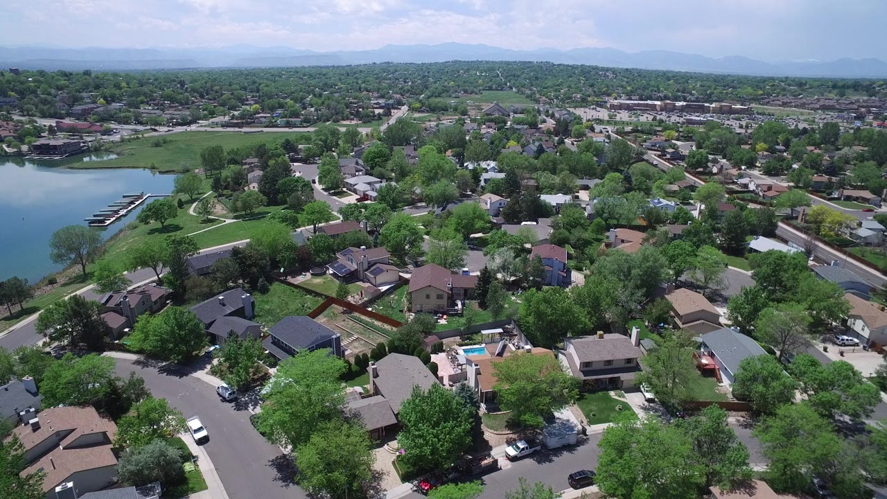 Aerial frame over Westminster, Colorado showing rooftops, fenced yards, green lawns, a lake edge with docks, and no visible people.