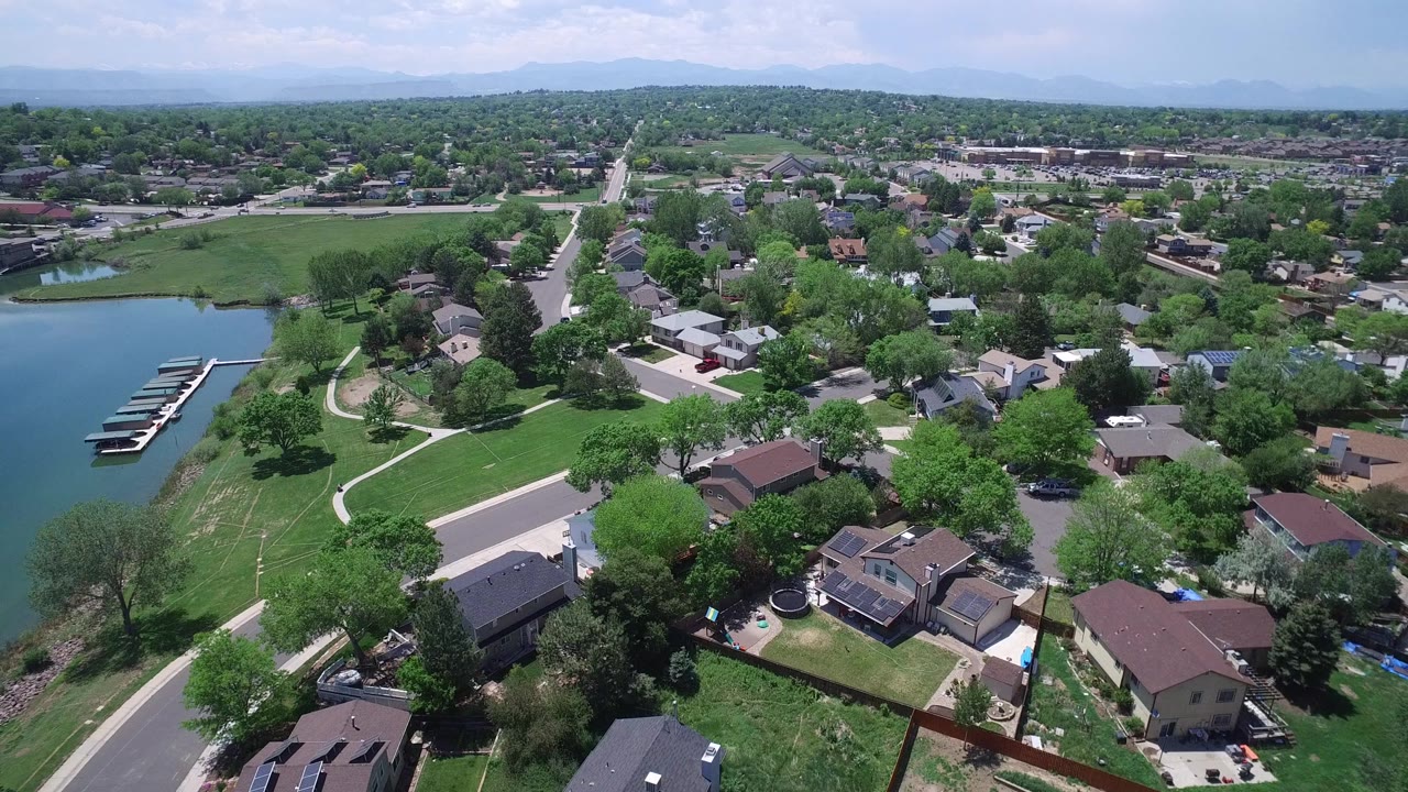 Aerial view in Westminster, Colorado with the lake and dock slips along the left edge, residential roofs in the foreground, open grass in the center, distant hills, and no visible people.