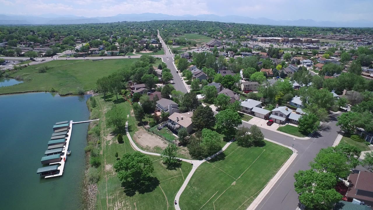 Aerial view in Westminster, Colorado showing an artificial lake with covered boat slips at left, green lawns and paths below, residential homes and roads beyond, and no visible people.