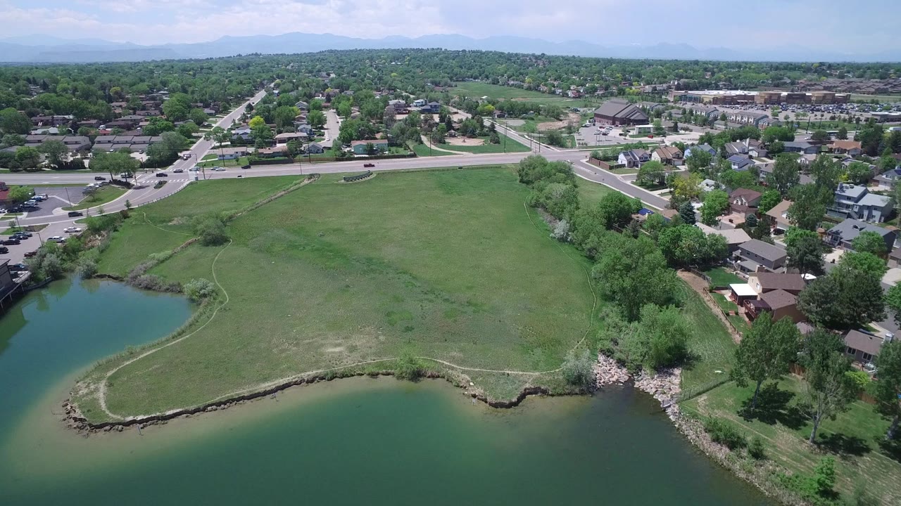 Aerial frame in Westminster, Colorado with the green pond shifted left, open grass centered, homes and roads behind it, and no visible people.