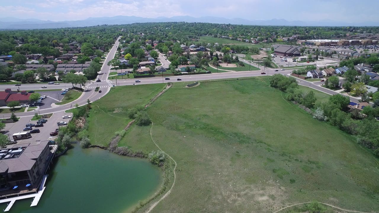 Aerial frame over Westminster, Colorado with a green pond at lower left, open grass across the center, roads with cars, distant homes, and no clearly visible people.