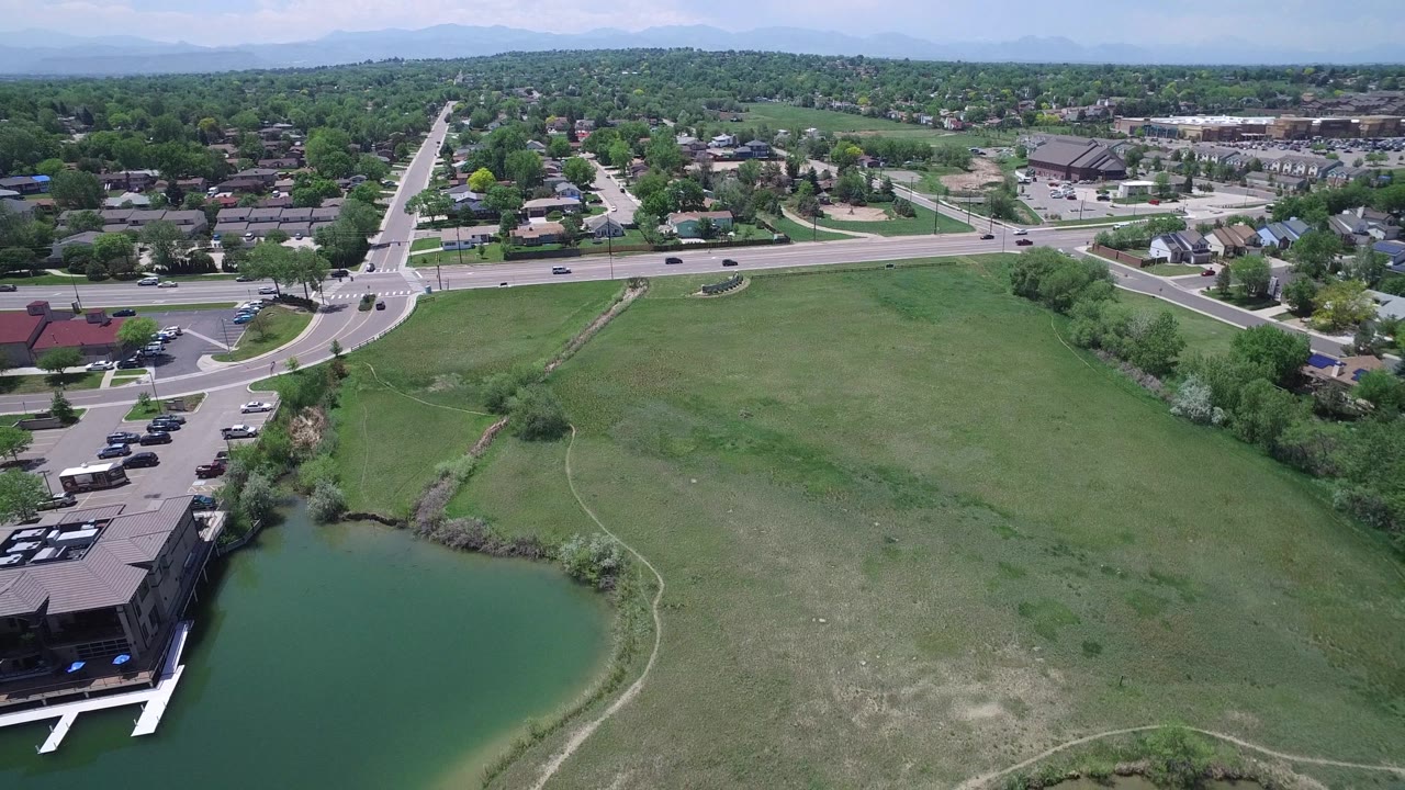 Aerial frame over Westminster, Colorado with the pond lower right, a broad grassy tract, a road intersection, moving traffic, dense tree cover, rooftops, and no identifiable people.