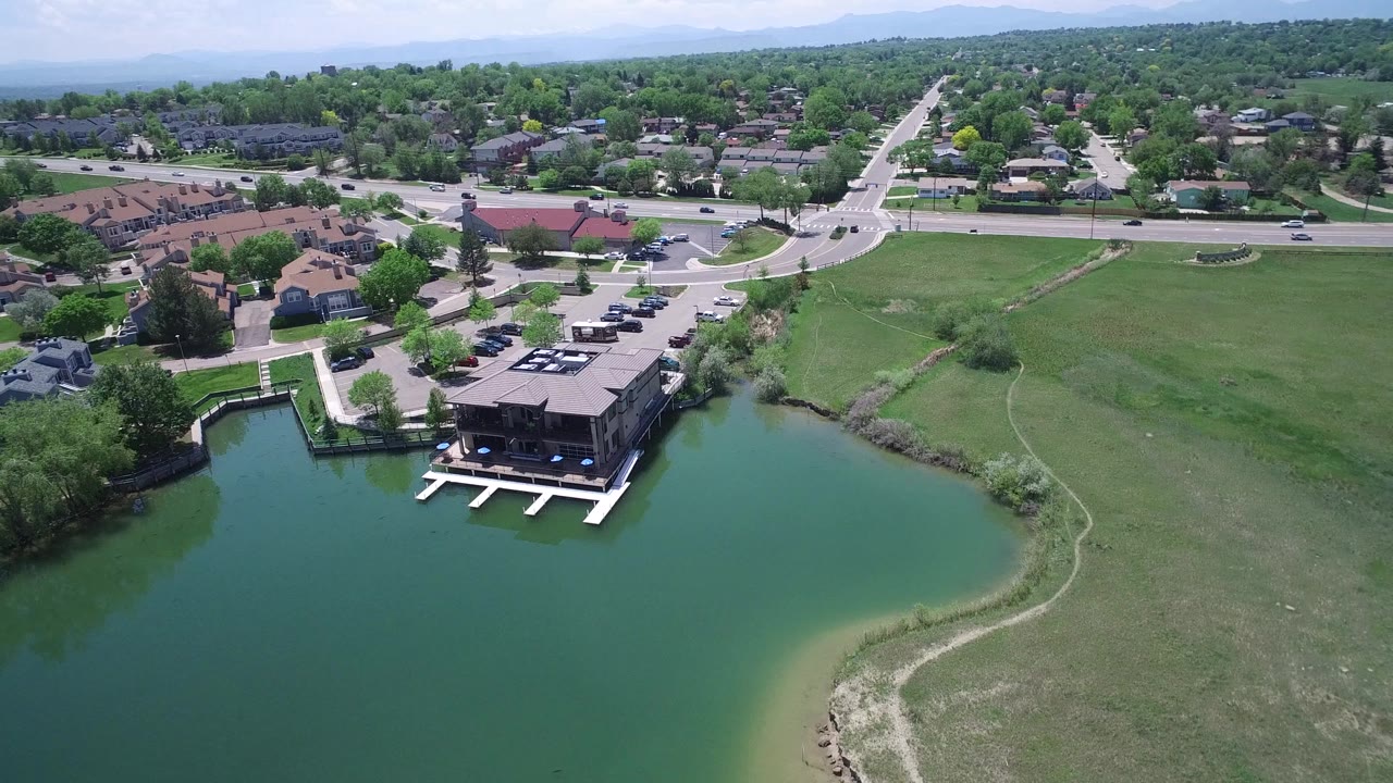 Aerial footage over Westminster, Colorado frames the lakefront building and docks near the lower center, with houses, parking areas, streets, trees, and no visible people beyond the water.