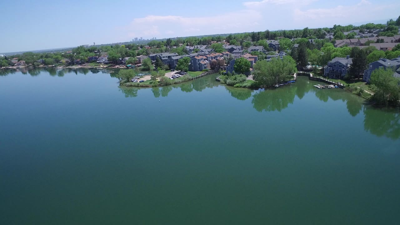Aerial frame of Westminster, Colorado lakefront homes and trees reflected faintly in still green-blue water, with no visible people.