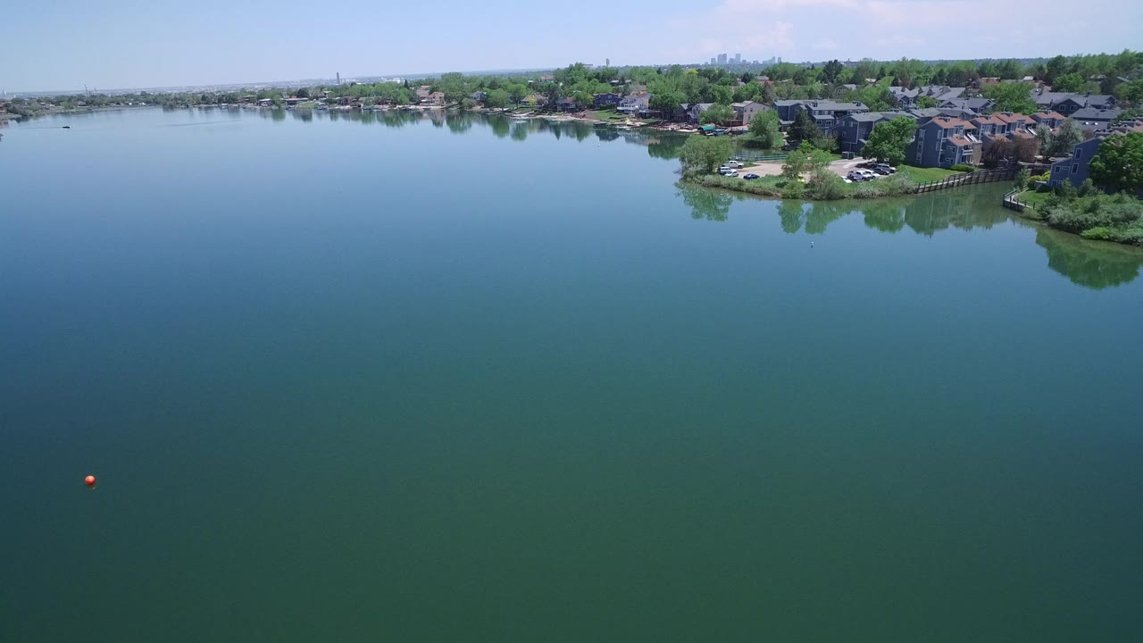 Aerial view over calm lake water with shoreline buildings in the distance.