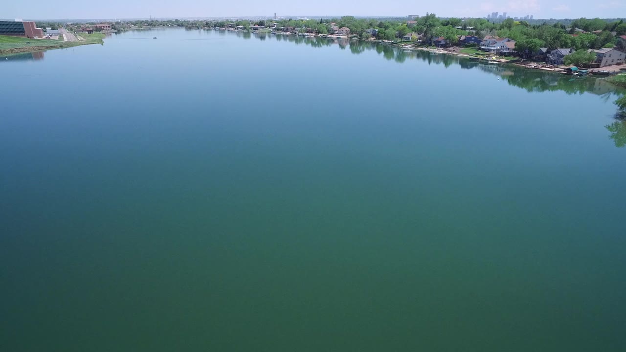Elevated view in Westminster, Colorado, looking across calm blue-green water toward shoreline homes, dense green trees, a pale sky, and no visible people.