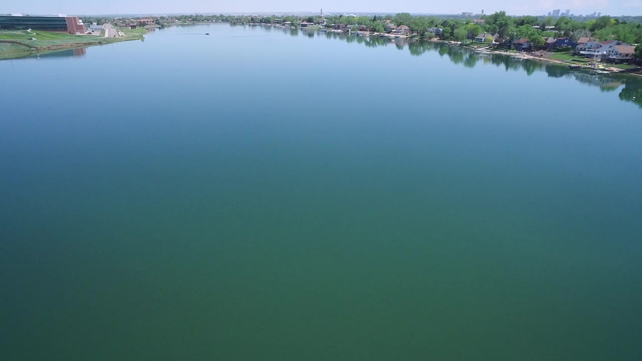 Wide aerial view of calm lake water in Westminster, Colorado, with the residential shoreline slightly farther across the upper frame and no visible people.