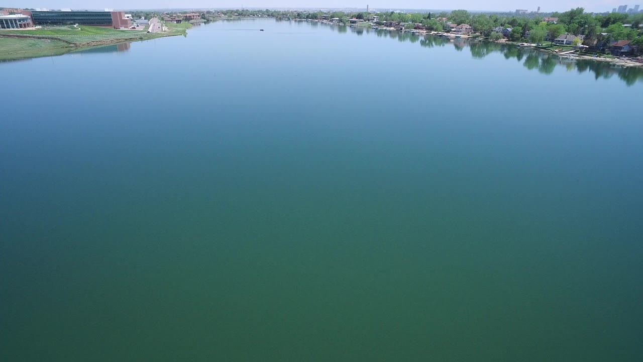 Aerial view over green-blue water with a distant tree-lined shore and buildings.