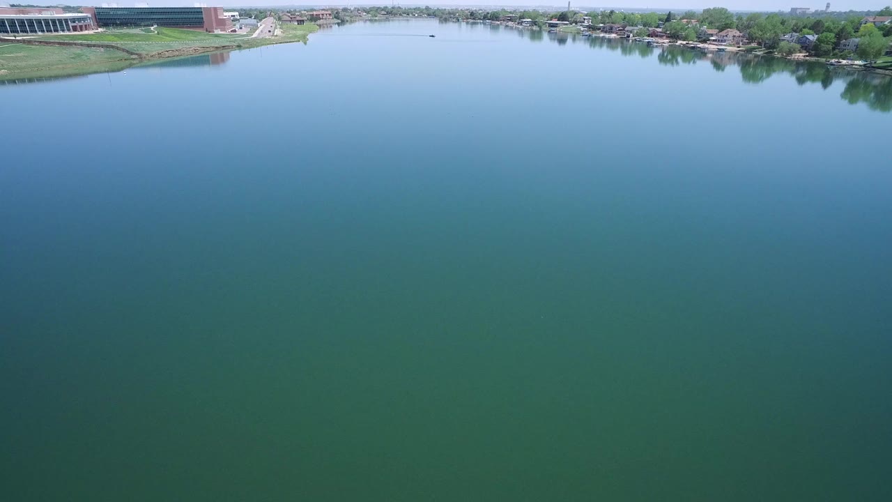 The second frame holds over the Westminster lake, showing a broad field of water with a distant line of homes, trees, shoreline lawns, and commercial structures near the top edge, no visible people.