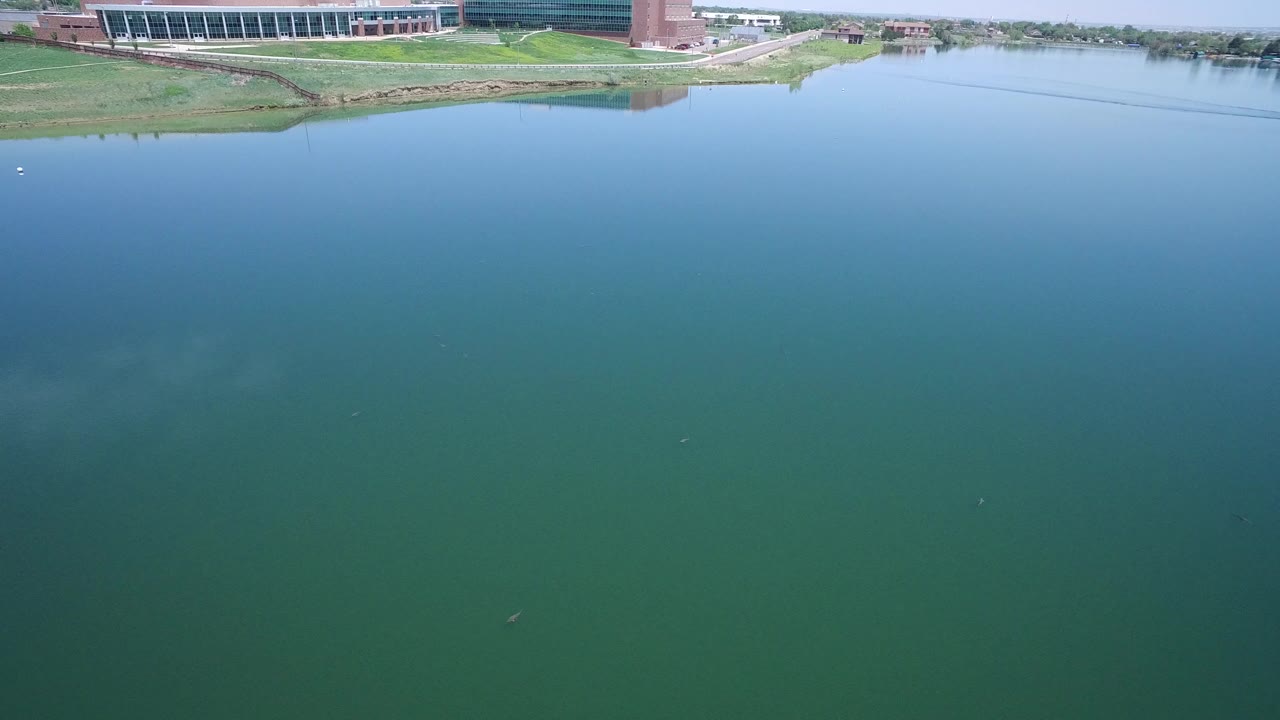 Opening frame over green-blue water in Westminster, Colorado, with grassy shoreline and modern buildings along the top edge, no visible people.