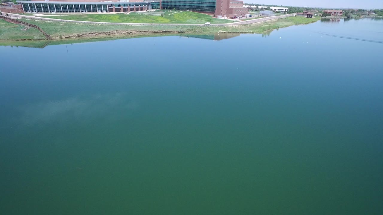 Opening frame of Westminster, Colorado aerial footage with broad green-blue water below, a grassy bank, road, path, and large modern buildings along the upper edge, with no visible people.