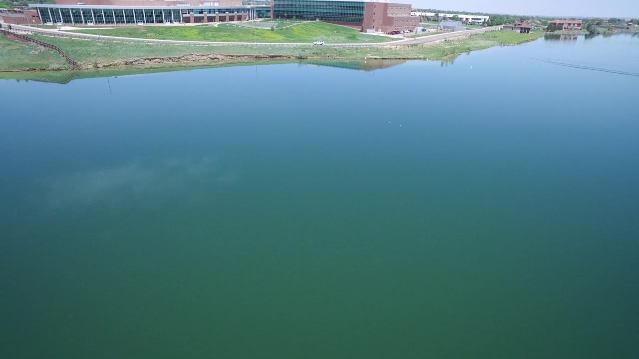 Aerial water view with modern buildings, road, and green bank along the far edge.