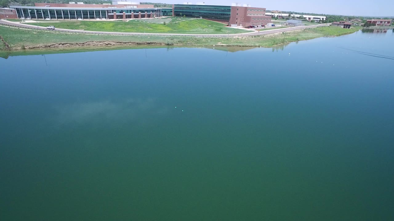 Aerial frame in Westminster, Colorado looking across blue-green water to a grassy shoreline and large brick and glass building, with no visible people.
