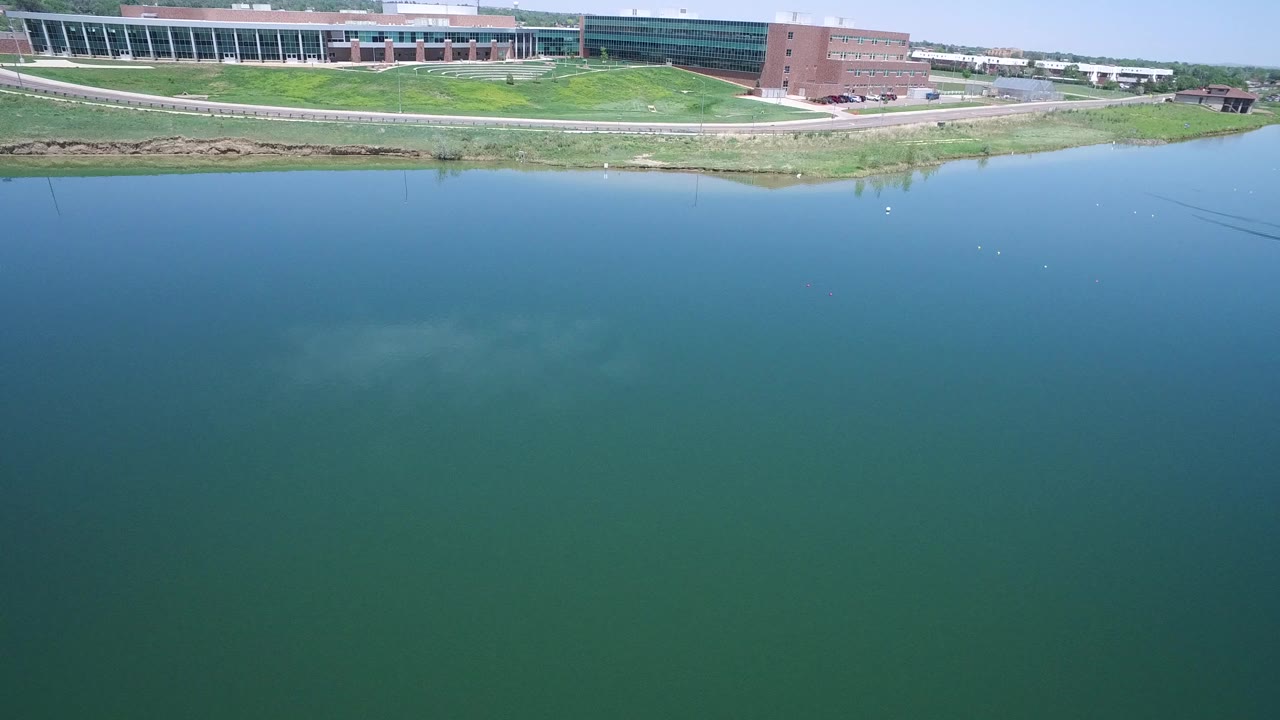 Aerial frame in Westminster, Colorado looking across blue-green water toward a large brick and glass building complex, grassy shoreline, and clear sky, with no visible people.