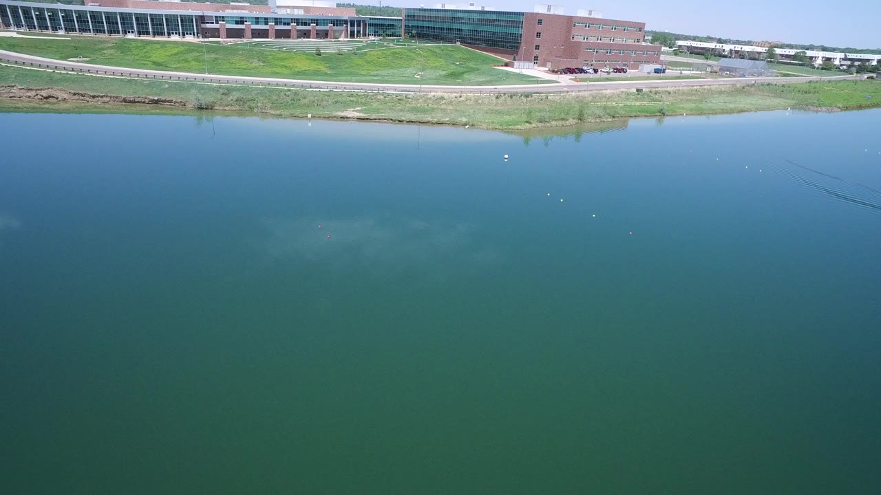 Aerial frame over Westminster, Colorado lake water with a grassy shoreline, paved path, and modern buildings across the top edge, with no visible people.