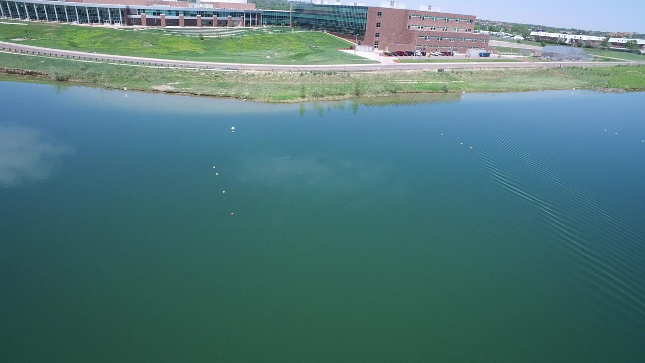Aerial view in Westminster, Colorado looking across blue-green water toward a grassy shore, road, and large buildings, with no visible people.