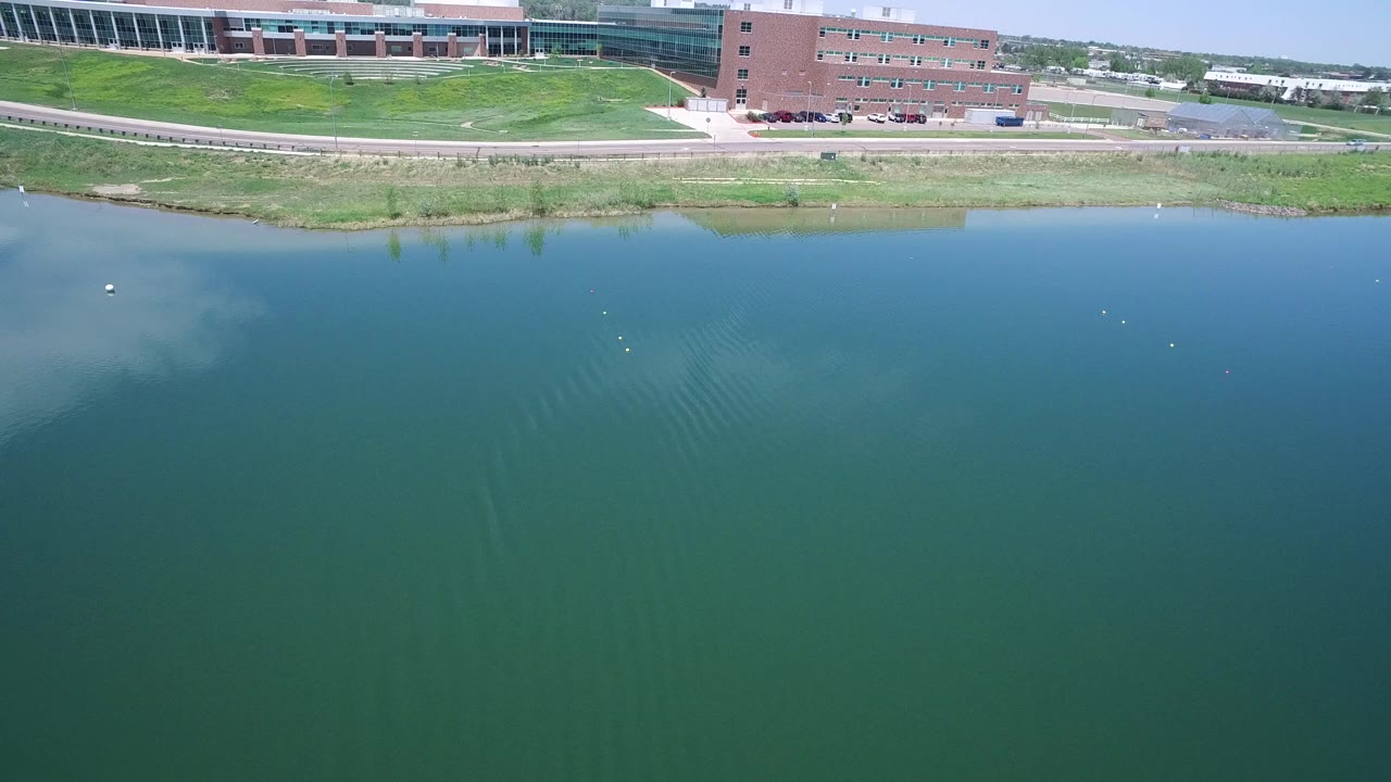 Drone view over a blue-green lake in Westminster, Colorado, with a brick building, grassy shoreline, road, parked cars, and no visible people.